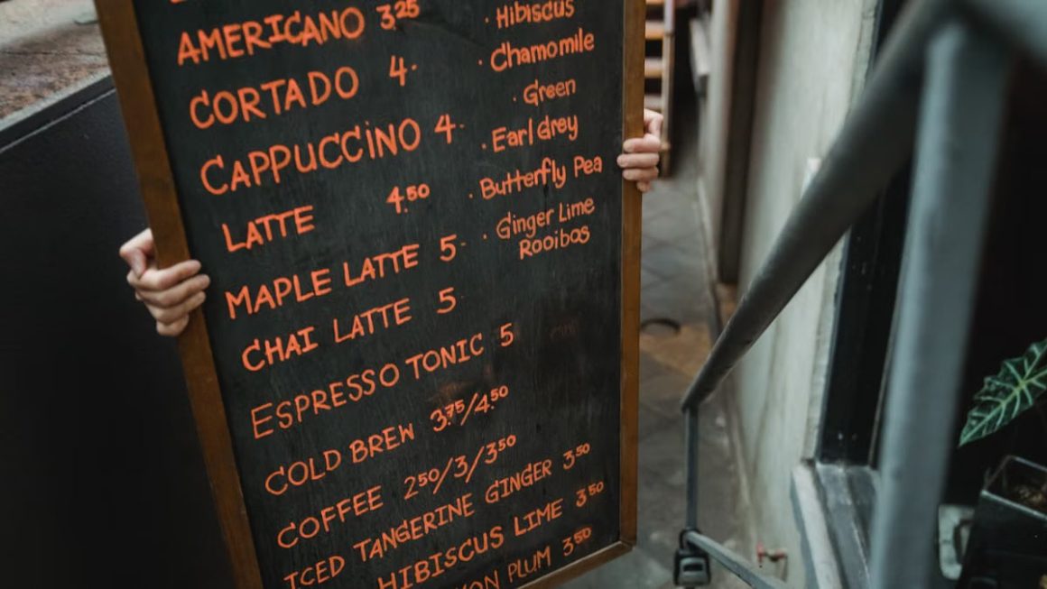 Close-up of a cafe menu on blackboard held by a person indoors, listing various coffee and tea options.